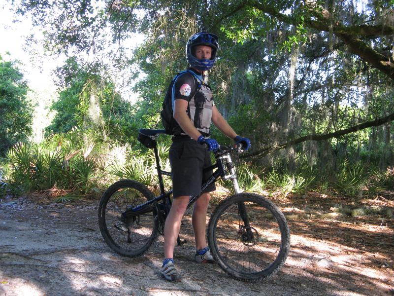 A mountain biker wearing a helmet and protective gear stands next to a black mountain bike. The background features a sun-dappled forest with green foliage and moss-covered trees, indicating an outdoor trail setting. Alafia River State Park mountain bike trail.