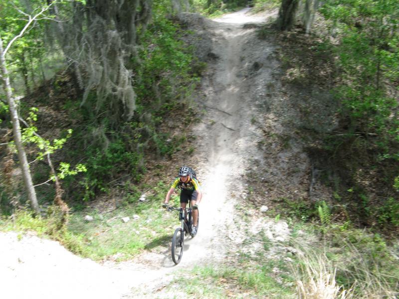 A person in a cycling outfit rides a mountain bike up a dirt trail surrounded by greenery and trees, with a steep incline in the background. Alafia River State Park mountain bike trail.