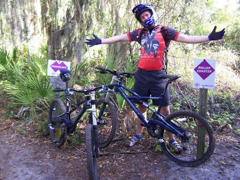 A person wearing a helmet and colorful biking apparel stands beside two mountain bikes on a trail. The background features lush greenery, and two signs can be seen—one labeled "Moonscape" and the other "Roller Coaster." The individual is smiling with arms outstretched, showcasing an excited attitude toward biking. Alafia River State Park mountain bike trail.