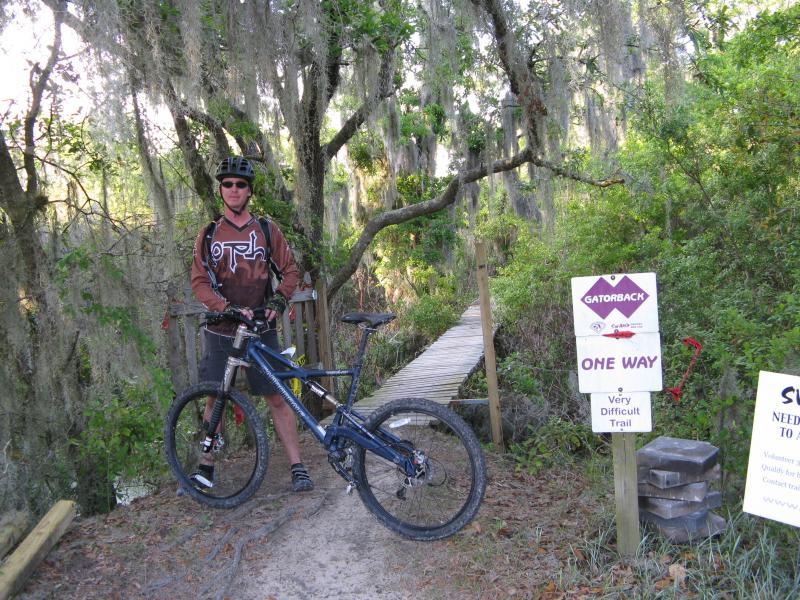 A mountain biker stands next to a blue bike on a narrow path, wearing a brown jersey and helmet. Behind him, a bridge leads into a wooded area, adorned with moss-covered trees. A sign nearby indicates a "Very Difficult Trail" and notes that it is a one-way path. Alafia River State Park mountain bike trail.