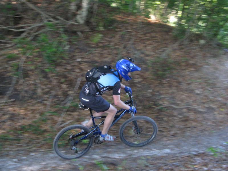 A mountain biker wearing a blue helmet and a backpack rides down a dirt trail surrounded by greenery. The bike is in motion, capturing a sense of speed and adventure. Alafia River State Park mountain bike trail.