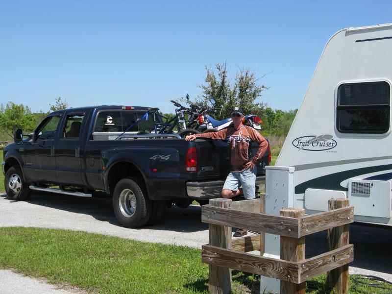 A man in casual clothing stands next to a black pickup truck parked beside a travel trailer. The truck is loaded with bicycles, and there is a wooden picnic table nearby. The scene is set on a sunny day with green grass and trees in the background. Alafia River State Park mountain bike trail.