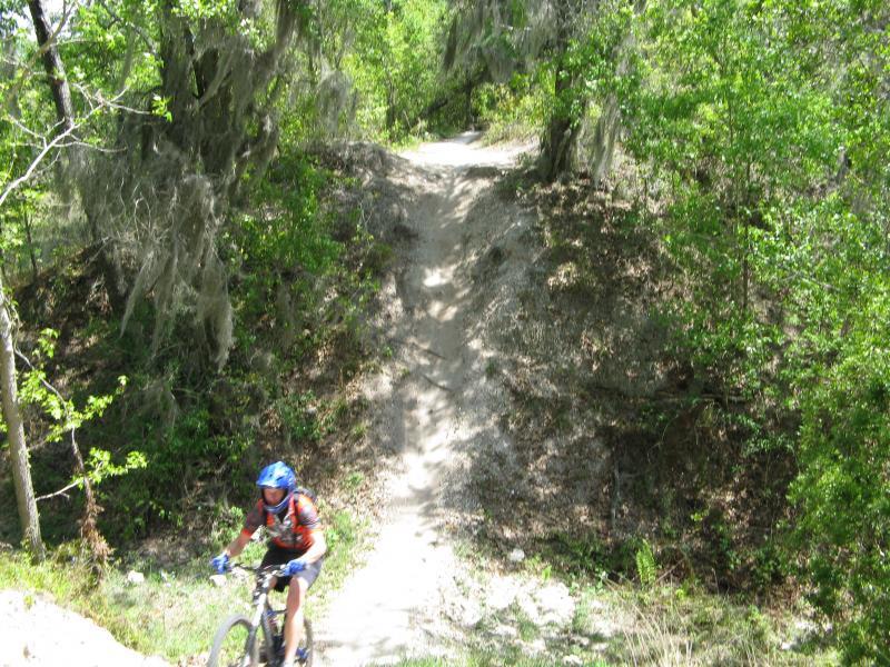 A mountain biker riding on a dirt trail through a lush, green forest with trees and Spanish moss in the background. The path features a hill, suggesting a challenging terrain for biking. Alafia River State Park mountain bike trail.