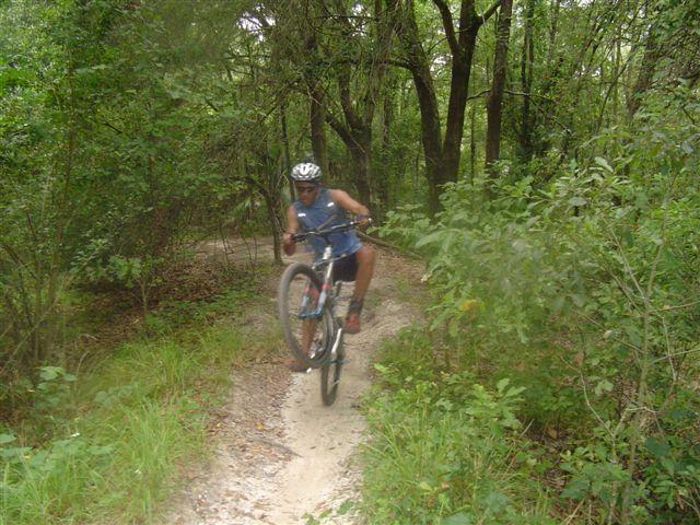 A mountain biker performing a wheelie on a dirt trail surrounded by green foliage and trees. The rider is wearing a helmet and is dressed in a tank top and shorts, showcasing an adventurous moment in nature. Alafia River State Park mountain bike trail.