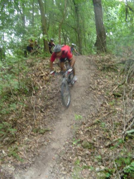 A mountain biker in a red shirt descends a dirt trail surrounded by trees and foliage, with a few onlookers visible in the background. The terrain appears rugged, suggesting an outdoor biking adventure. Alafia River State Park mountain bike trail.