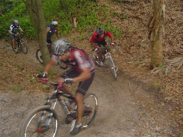 Four mountain bikers navigate a dirt trail surrounded by trees and foliage. The lead rider is in motion, leaning forward on his bike, while the others follow closely behind. The scene captures the excitement and intensity of mountain biking in a natural setting. Alafia River State Park mountain bike trail.
