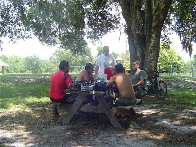 Five men gathered around a picnic table in a shaded park area, engaged in conversation. Some of them are shirtless, wearing shorts, and one person is in a white t-shirt with an apron. A bicycle is seen nearby. The scene features lush greenery and a playground in the background, suggesting a casual outdoor setting. Alafia River State Park mountain bike trail.