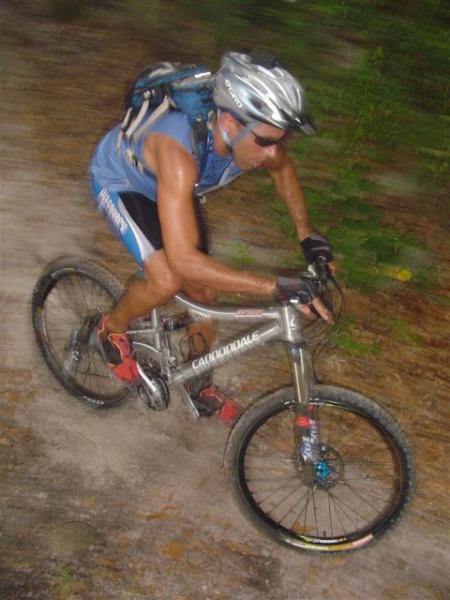 A mountain biker navigating a dirt trail, wearing a helmet and sunglasses, with a backpack. The rider is captured mid-motion, demonstrating an active and dynamic posture on a silver mountain bike. The background features blurred greenery, indicating speed and movement. Alafia River State Park mountain bike trail.