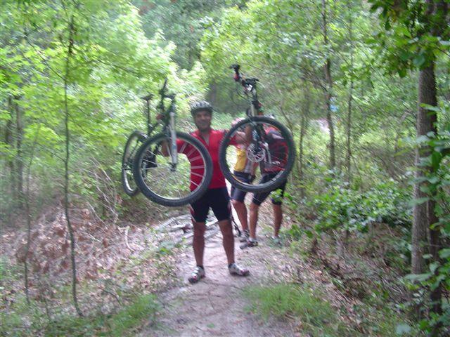 A person in a red cycling jersey and helmet is holding two mountain bikes above their head while standing on a wooded trail. Another cyclist in a yellow shirt is visible in the background, navigating the path surrounded by greenery. Alafia River State Park mountain bike trail.