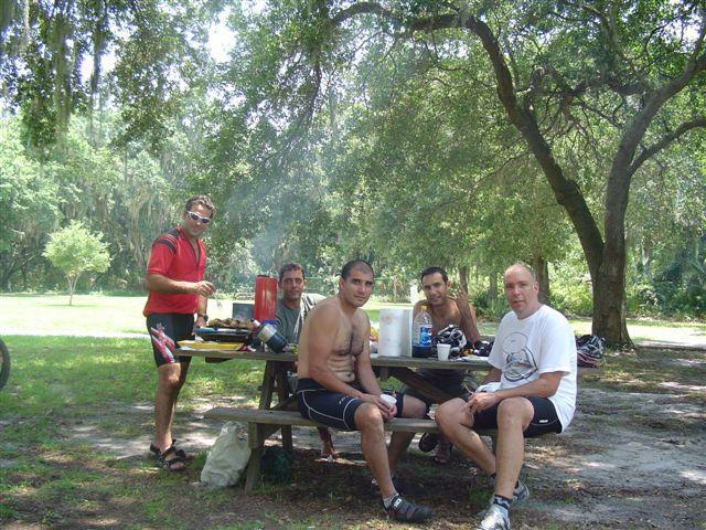 A group of five men enjoying a casual outdoor gathering around a picnic table. They are surrounded by trees in a sunny park setting, with some cooking equipment and drinks on the table. The atmosphere appears relaxed and social, with one man cooking while the others sit and chat. Alafia River State Park mountain bike trail.