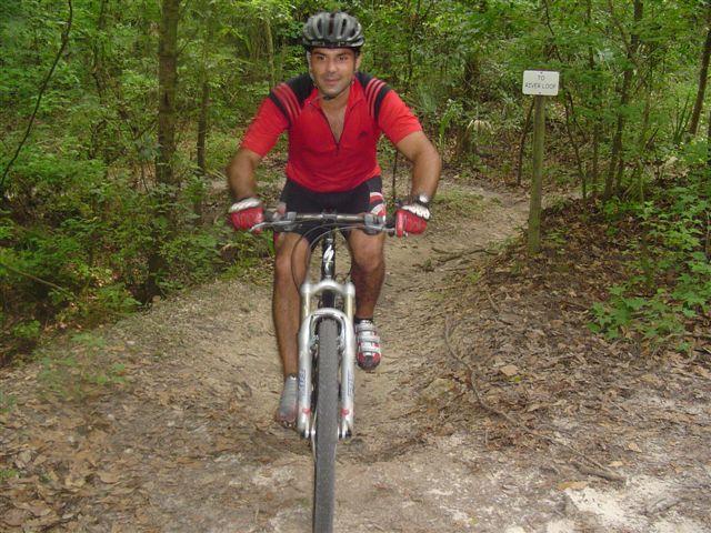 A man wearing a helmet and a red cycling jersey rides a mountain bike on a dirt trail surrounded by lush green trees. A sign indicating a trailhead can be seen in the background. The scene captures the spirit of outdoor adventure and mountain biking. Alafia River State Park mountain bike trail.