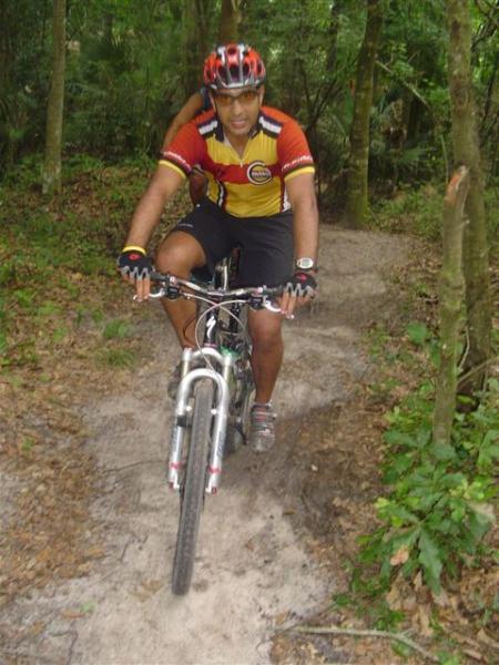 A mountain biker riding on a dirt path through a lush, green forest. The cyclist is wearing a colorful jersey and helmet, with a focused expression, demonstrating balance and skill as he navigates the trail. Alafia River State Park mountain bike trail.
