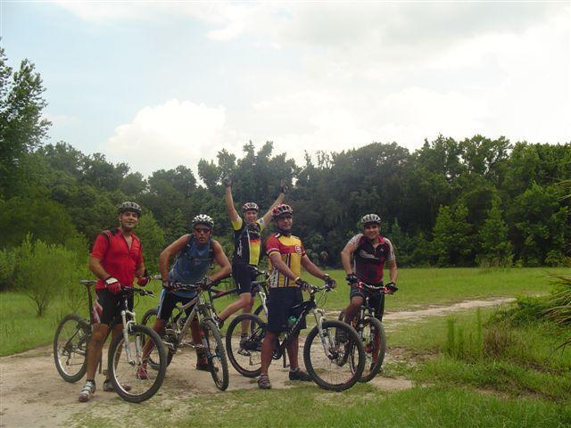 A group of five mountain bikers posing on a dirt path surrounded by greenery. They are wearing cycling attire and helmets, with their bikes parked beside them. The atmosphere is casual and friendly, with trees and a cloudy sky in the background. Alafia River State Park mountain bike trail.