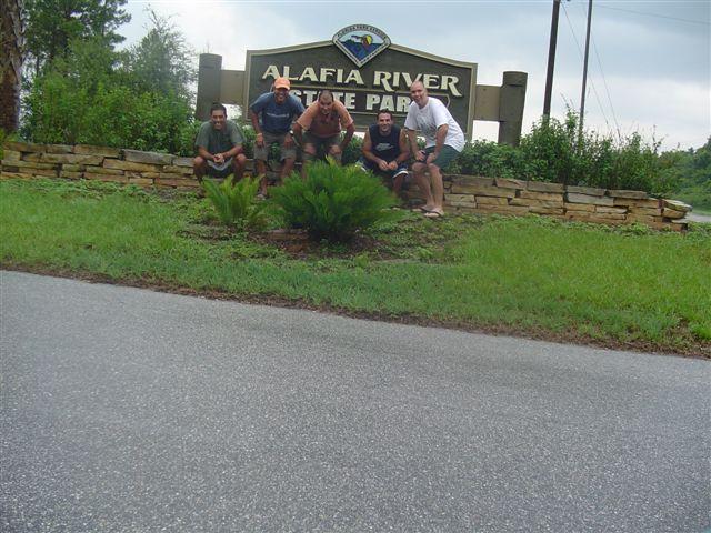 A group of four men posing in front of the Alafia River State Park sign, surrounded by lush greenery and landscaping. The men are dressed casually, with smiles on their faces, capturing a moment of camaraderie at the park entrance. The sky is overcast, indicating a cloudy day. Alafia River State Park mountain bike trail.