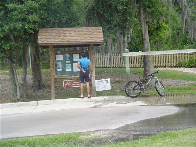A person in athletic clothing stands in front of an informational sign at a park, reading the posted notices. A bicycle leans against a nearby post, and there are trees and a picket fence in the background. The ground is wet, suggesting recent rain. Alafia River State Park mountain bike trail.
