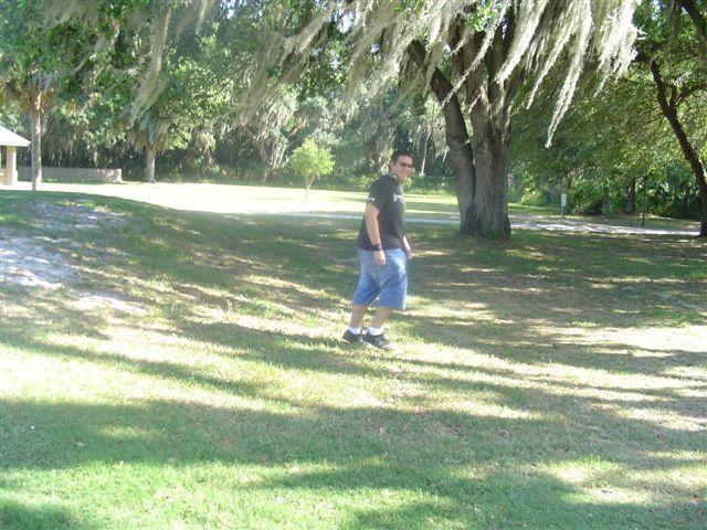 A person walking on grassy terrain in a park, surrounded by trees with Spanish moss hanging from the branches. The sun is shining, creating a bright and natural atmosphere. Alafia River State Park mountain bike trail.