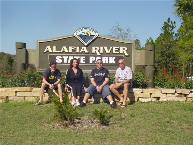 Four people sit in front of the Alafia River State Park sign, with greenery and trees in the background. They appear to be enjoying a sunny day outdoors. The sign prominently displays the name of the park. Alafia River State Park mountain bike trail.
