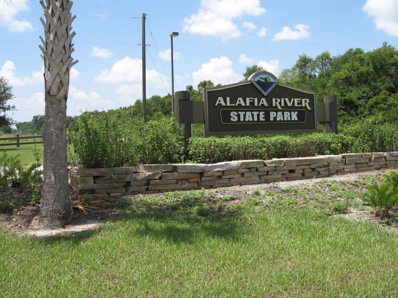 Sign for Alafia River State Park, featuring green lettering on a dark sign. The sign is surrounded by lush greenery, a stone wall, and a palm tree in the foreground, under a partly cloudy blue sky. Alafia River State Park mountain bike trail.