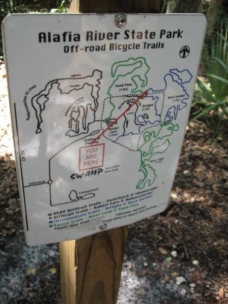 Map of Alafia River State Park showcasing off-road bicycle trails. The sign indicates various bike paths in different colors, with a highlighted area marked "YOU ARE HERE" near a region labeled "SWAMP." Trail difficulty levels are also noted on the sign. Alafia River State Park mountain bike trail.