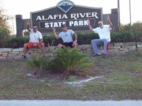 Three individuals sitting on a stone wall in front of the Alafia River State Park sign, all smiling and making the number one gesture with their hands. The background features greenery and a clear sky, indicating a sunny day. Alafia River State Park mountain bike trail.