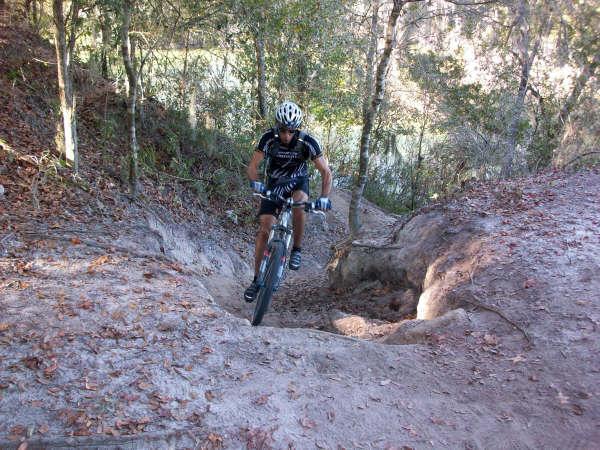 A mountain biker navigating a steep, sandy trail surrounded by trees, with a sunny background and a river visible in the distance. Alafia River State Park mountain bike trail.