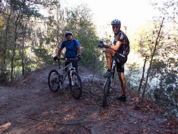 Two mountain bikers pause on a dirt trail surrounded by trees. One rider is sitting on their bike with one leg raised, while the other stands next to a white mountain bike. The scene is set in a natural, wooded area, suggesting a moment of rest during their biking adventure. Alafia River State Park mountain bike trail.