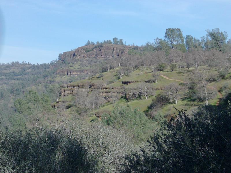 A scenic view of a rugged hillside with sparse trees, showcasing sloped terrain and rocky outcrops under a clear blue sky. The landscape features a mix of greenery and dry vegetation, highlighting the natural contours of the land. Upper Bidwell Park mountain bike trail.