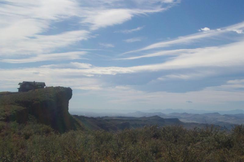 A scenic view of a high cliff with a small wooden house perched on top, surrounded by lush green vegetation and a vast landscape of rolling hills and mountains in the distance under a blue sky with scattered clouds. Coyote Ridge mountain bike trail.