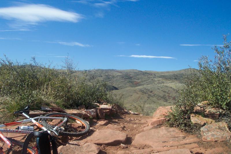 A mountain biking scene showcasing a trail with rocky terrain, featuring a bicycle lying on the ground. In the background, rolling hills are visible under a clear blue sky with a few wispy clouds. Scrubby vegetation surrounds the area, indicating a natural outdoor setting. Coyote Ridge mountain bike trail.