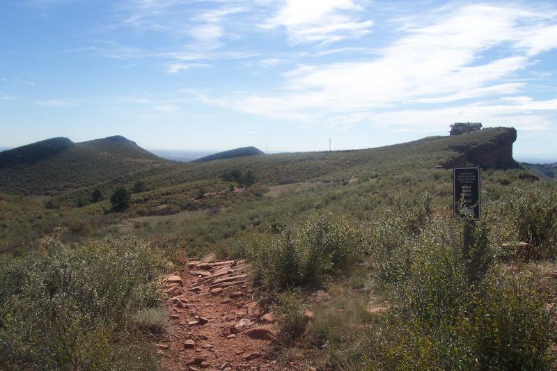 A scenic view of rolling hills and rocky terrain under a blue sky, featuring a hiking path leading toward a distant rocky outcrop with a small structure on top. Lush green vegetation and shrubs are scattered throughout the landscape. A trail sign can be seen along the path, indicating the area for hikers. Coyote Ridge mountain bike trail.