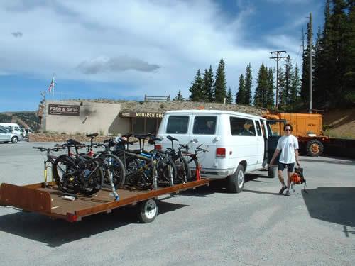 A man walking next to a white van with a trailer carrying several mountain bikes parked in a rest area surrounded by trees. In the background, there is a building with a sign for food and gifts. Monarch Crest Trail mountain bike trail.