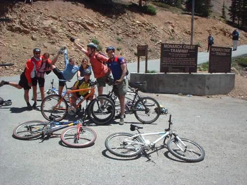 A group of six mountain bikers joyfully posing with their bikes at the Monarch Crest Tramway. Some are standing, while one person is playfully kicking their leg up. Several bikes are scattered on the ground nearby, and a sign indicating the tramway is visible in the background. The setting is outdoors, with rocky terrain and trees in the background. Monarch Crest Trail mountain bike trail.