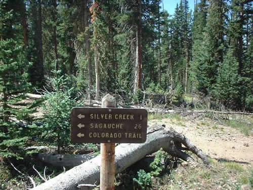 Signpost indicating trail directions in a forested area, with arrows pointing left towards Silver Creek (1 mile) and Sagauche (26 miles), and listing the Colorado Trail. Surrounding the sign are tall trees and natural vegetation. Monarch Crest Trail mountain bike trail.