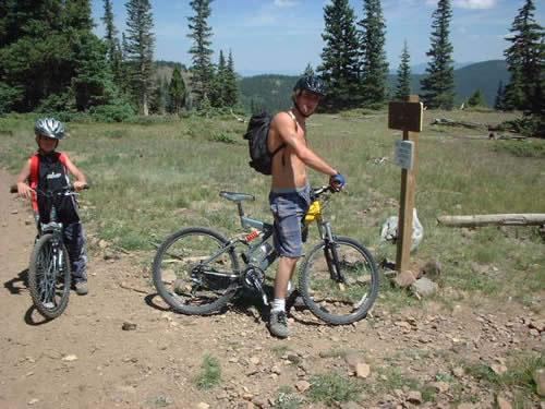 A young man with a backpack and no shirt stands beside his mountain bike on a dirt trail, looking at a trail sign. A child in a black sleeveless shirt and helmet is positioned nearby with another bike. The scene is set in a lush, mountainous area with tall trees and clear blue skies in the background. Monarch Crest Trail mountain bike trail.
