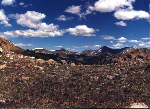 A panoramic view of a mountainous landscape under a blue sky, with fluffy white clouds. The scene features rugged mountain peaks in the background, some with patches of snow, and a foreground of rocky terrain with sparse vegetation. Monarch Crest Trail mountain bike trail.