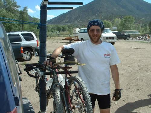 A man wearing a bandana and a white t-shirt stands beside a blue vehicle, holding onto a bicycle rack that is mounted on the back. The background features mountains and several vehicles parked in a recreational area. The setting appears to be outdoors, likely at a biking or camping site. Monarch Crest Trail mountain bike trail.