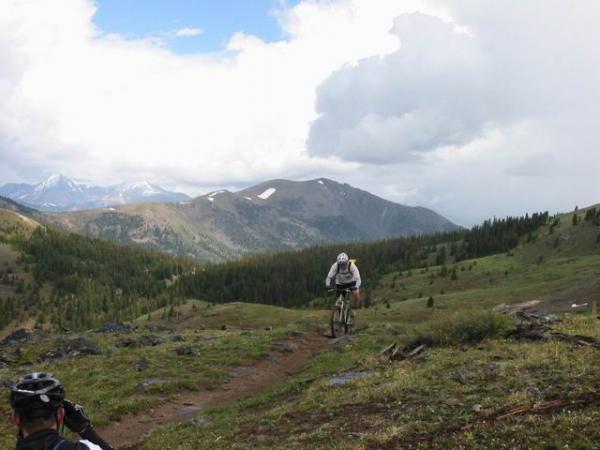 A mountain biker rides along a dirt trail through a lush, green landscape with mountains in the background under partly cloudy skies. The scene showcases the natural beauty of the area, with a mix of forested hills and rocky terrain. Monarch Crest Trail mountain bike trail.