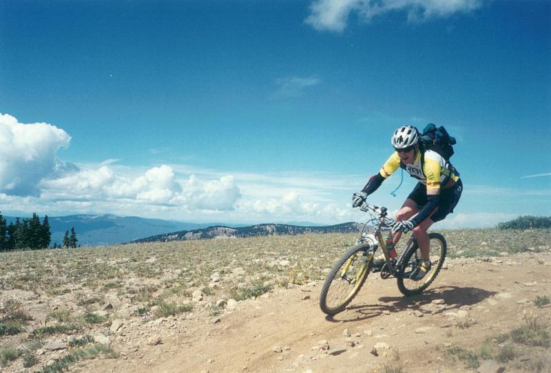 A mountain biker rides along a rocky trail under a blue sky with fluffy clouds. The cyclist, wearing a helmet and a yellow jersey, leans forward on the bike as they navigate the uneven terrain, with green trees and distant mountains visible in the background. Monarch Crest Trail mountain bike trail.