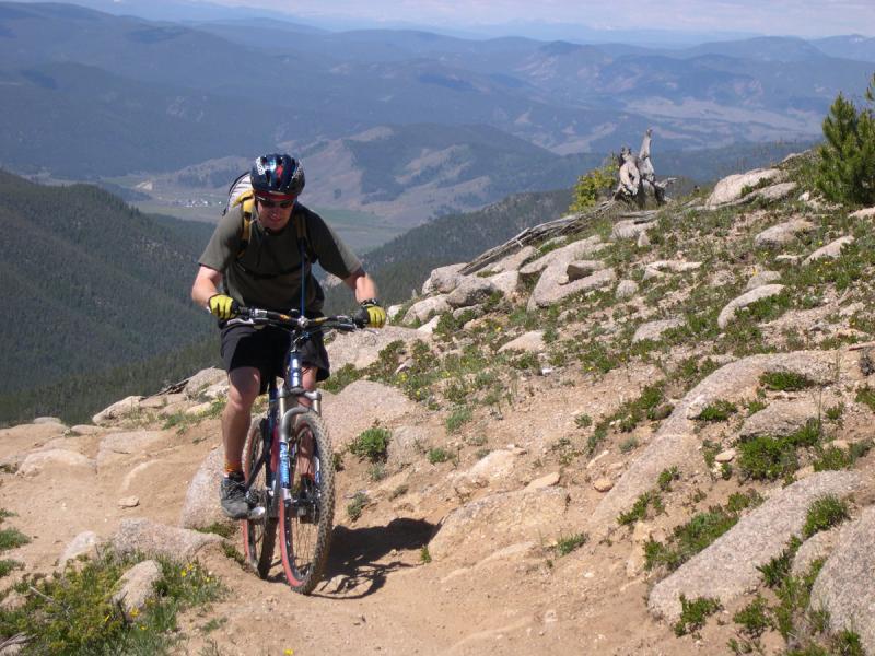 A mountain biker ascends a rocky trail on a hillside, surrounded by green vegetation and a scenic mountainous landscape in the background. The cyclist is wearing a helmet and gloves, and is focused on navigating the terrain. The sky is clear, and the view extends into the distance, showcasing rolling hills and valleys. Monarch Crest Trail mountain bike trail.