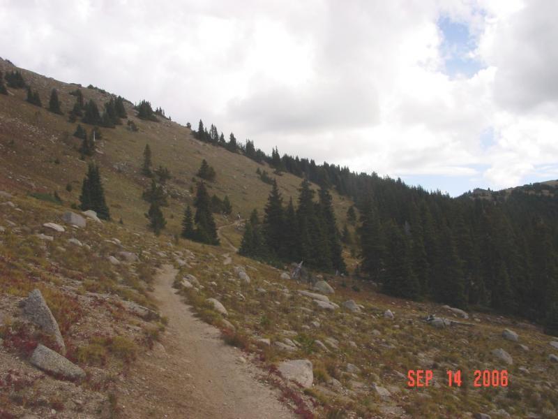 A winding dirt path leads through a mountainous landscape, with rocky terrain and patches of grass. Tall evergreen trees line the hillside, under a cloudy sky. The date in the bottom right corner reads September 14, 2006. Monarch Crest Trail mountain bike trail.