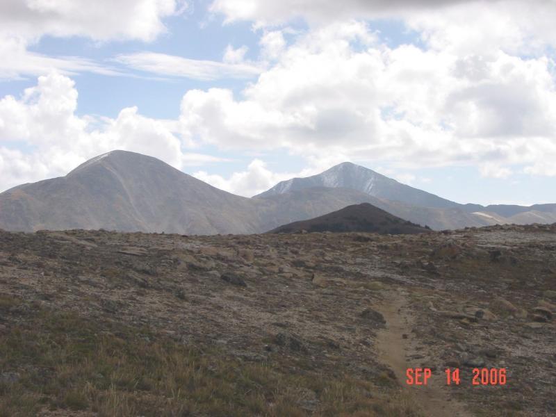 A scenic view of a mountainous landscape with two prominent peaks in the background. The foreground features a rocky terrain with a visible dirt path leading into the distance. The sky is partly cloudy with patches of blue, and the peaks are partially covered in snow. The scene conveys a sense of natural beauty and tranquility. The date "SEP 14 2006" is displayed in the lower right corner. Monarch Crest Trail mountain bike trail.