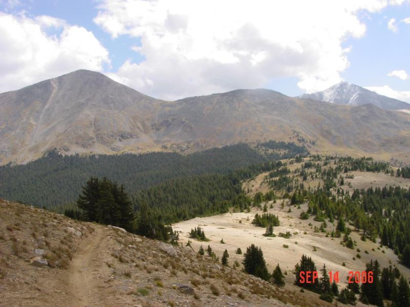 A scenic view of mountains under a partly cloudy sky, featuring a trail on the left leading through hilly terrain. The foreground shows a mix of rocky ground and patches of grass with scattered trees, while the background displays towering mountains with varying shades of brown and green. Monarch Crest Trail mountain bike trail.