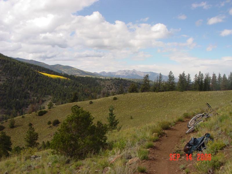A scenic landscape featuring rolling hills, greenery, and distant mountains under a partly cloudy sky. A dirt path runs through the foreground, with a bicycle and a piece of clothing resting nearby, suggesting outdoor activities like biking or hiking in a natural setting. Monarch Crest Trail mountain bike trail.