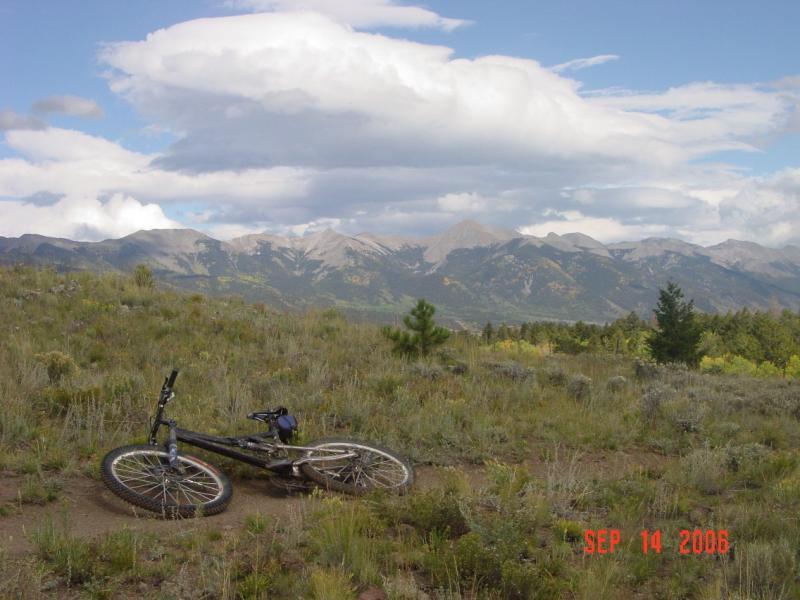 A mountain bike lying on a grassy area with mountains and a cloudy sky in the background. The scene captures the tranquil beauty of nature, showcasing the rugged terrain and distant peaks. The date in the corner indicates that the photo was taken on September 14, 2006. Monarch Crest Trail mountain bike trail.