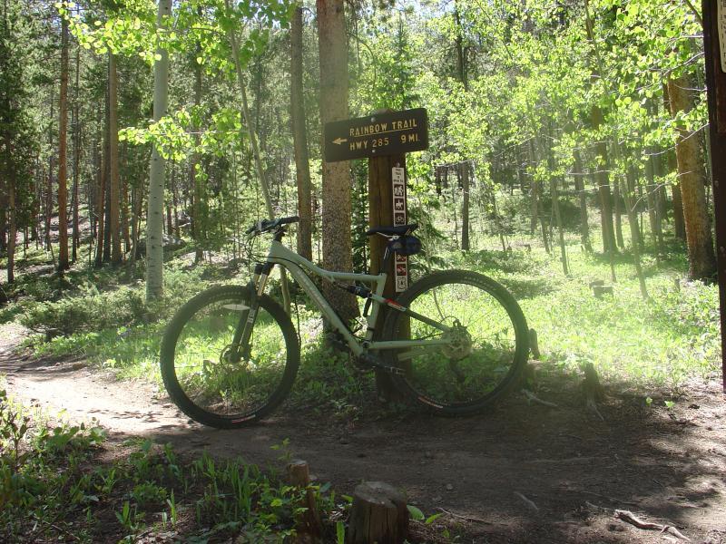 A mountain bike leaning against a sign for the Rainbow Trail, indicating a distance of 9 miles to Highway 285, surrounded by lush green trees and sunlight filtering through the foliage. Monarch Crest Trail mountain bike trail.