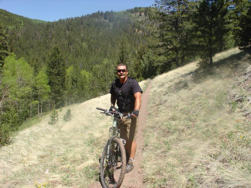 A mountain biker posing on a dirt trail surrounded by lush green trees and rolling hills, with a sunny blue sky in the background. The rider is wearing a black t-shirt and shorts, smiling while holding onto his bicycle. Monarch Crest Trail mountain bike trail.