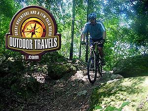 A mountain biker navigating a rocky trail in a lush green forest, with a logo for "Outdoor Travels" featuring a compass and the tagline "Great Adventure and a Few Cold Beers." Santos mountain bike trail.