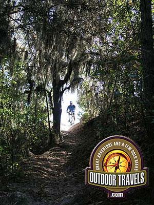 A cyclist riding on a narrow trail surrounded by lush greenery and trees, with sunlight filtering through the leaves. The image features the logo of Outdoor Travels in the corner, promoting outdoor adventures. Balm Boyette Scrub Preserve mountain bike trail.