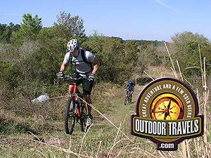 A person riding a mountain bike on a grassy trail surrounded by trees and vegetation, with another cyclist in the background. An "Outdoor Travels" logo is displayed in the lower right corner. Balm Boyette Scrub Preserve mountain bike trail.