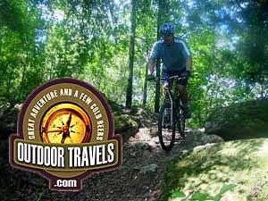 A person biking on a wooded trail, surrounded by lush green trees and rocks. The image features a logo for "Outdoor Travels," emphasizing adventure and outdoor experiences. Santos mountain bike trail.